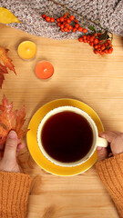 female hands holding a cup of hot tea or coffee, autumn flat in Scandinavian style, with yellow leaves, cozy knitwear, candles, a blank for the designer, mock up, copy space