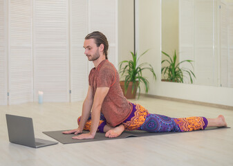 A young male yogi practices and teaches yoga online on a laptop in the Studio, performing and...
