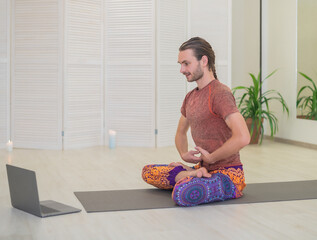 A young male yogi practices and teaches yoga online on a laptop in the Studio, performing and...