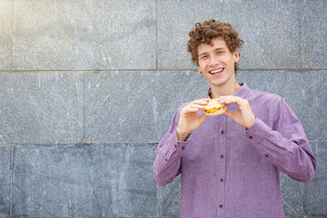 Takeaway, fast food and people concept - happy smiling European young curly man, wearing casual clothes, holding small hamburger and looking at camera on gray background. Empty copy space.