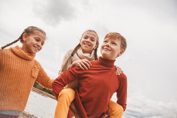 Preteen kids, boy and twin girls hugging in fall park with lake.  Friends wearing warm knit sweaters
