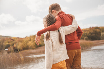 back view of preteen kids, boy and girl hugging in fall park with lake.  Friends wearing warm knit sweaters
