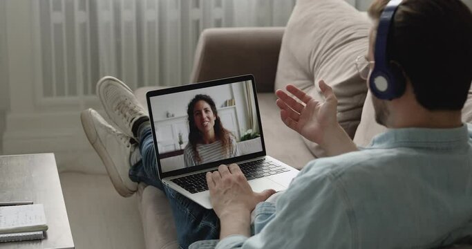 Relaxed man resting on couch wearing headphones talking with woman use videocall app, pc screen view over male shoulder. Couple in separation long distance relationship, video conference usage concept