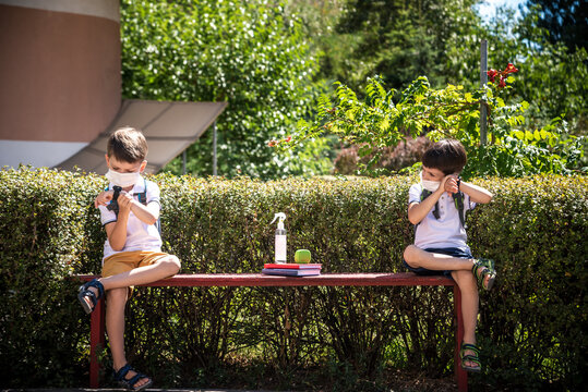 Coronavirus school reopening concept: a school boys sit on bench in front of a school wearing a face mask. Communicating trough smart watch. Covid-19, health, education, safety, and back to school