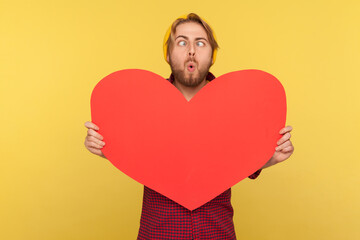 Funny romantic guy holding big read heart and looking at camera cross-eyed, having fun, demonstrating love feelings with huge greeting card on Valentines Day. studio shot isolated on yellow background