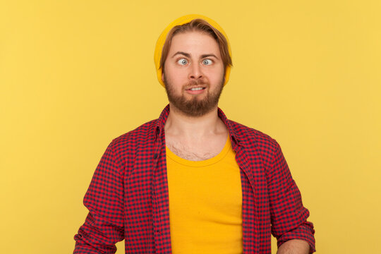 Funny dumb hipster bearded guy in beanie hat and checkered shirt making silly face with eyes crossed, looking at camera stupid brainless expression. indoor studio shot isolated on yellow background