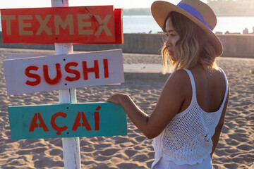 Beautiful asian woman standing and relaxing on the beach at sunset next to the wooden sign (in english: Sushi, Berries and Coffee). Vietnamese girl having holiday in a luxury resort. Lifestyle concept