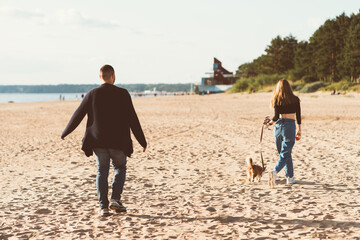 Back view of young happy couple and dog walking on beach. Handsome man and beautiful woman holding Corgi puppy on leash. Walking in summer nature of people in love