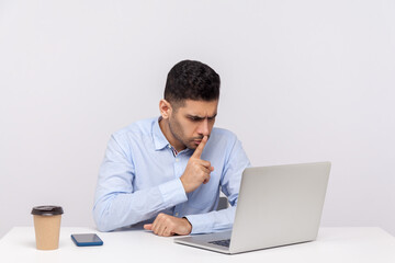Please, be quiet! Strict bossy man sitting office workplace, asking for silence while talking video call on laptop, keeping finger on lips and hushing. indoor studio shot isolated on white background