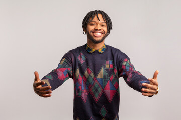 Friendly cheerful african guy with dreadlocks in casual style sweatshirt going to hug with toothy smile on face, happy to see, hospitality. Indoor studio shot isolated on gray background
