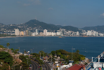 Fototapeta na wymiar Acapulco, Mexico - November 25, 2008: The blue water bay with beach and highrise buildings along shoreline. Backdrop of green hills loaded with houses under blue sky. Front is freeway. 