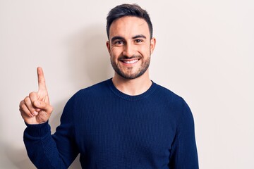 Young handsome man with beard wearing casual sweater standing over white background smiling with an idea or question pointing finger up with happy face, number one