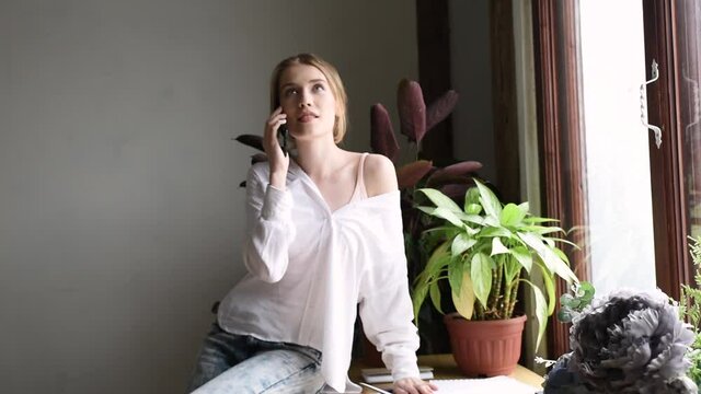 Happy woman in white blouse and blue jeans speaks on the phone at home office. Table by the window, green plants.