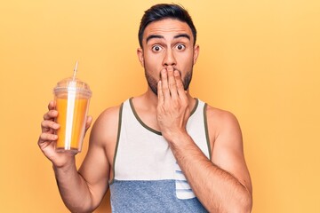 Handsome man with beard wearing sleeveless t-shirt drinking glass of healthy orange juice covering mouth with hand, shocked and afraid for mistake. Surprised expression