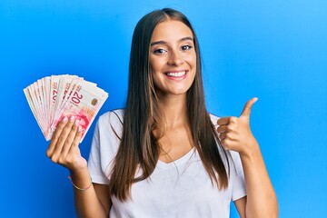 Young hispanic woman holding israel shekels smiling happy and positive, thumb up doing excellent and approval sign