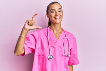 Young hispanic woman wearing doctor uniform and stethoscope smiling and confident gesturing with hand doing small size sign with fingers looking and the camera. measure concept.