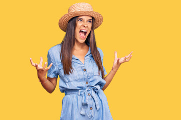 Young hispanic woman wearing summer hat crazy and mad shouting and yelling with aggressive expression and arms raised. frustration concept.