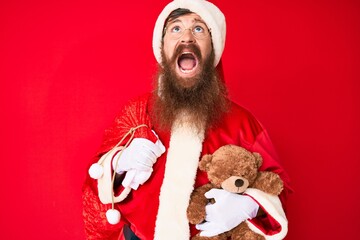Handsome young red head man with long beard wearing santa claus costume holding teddy bear angry and mad screaming frustrated and furious, shouting with anger looking up.