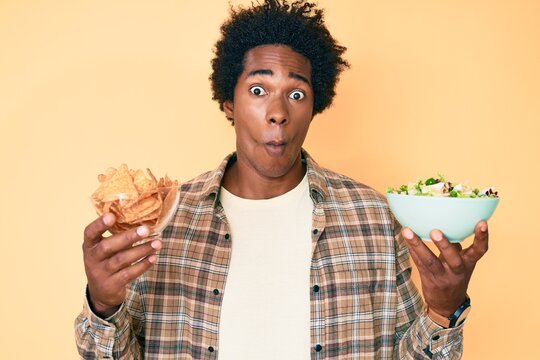 Handsome African American Man With Afro Hair Holding Nachos And Healthy Salad Making Fish Face With Mouth And Squinting Eyes, Crazy And Comical.