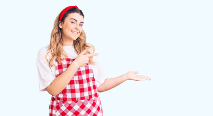 Young caucasian woman wearing apron amazed and smiling to the camera while presenting with hand and pointing with finger.