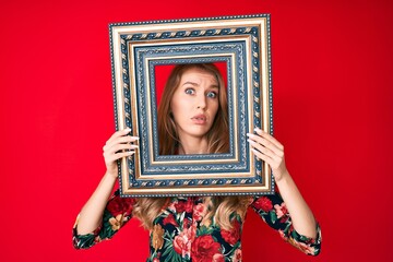 Young caucasian woman with blond hair holding empty frame clueless and confused expression. doubt concept.