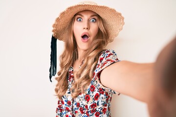 Young caucasian woman with blond hair wearing summer hat taking a selfie scared and amazed with open mouth for surprise, disbelief face