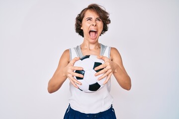 Young hispanic woman holding soccer ball angry and mad screaming frustrated and furious, shouting with anger looking up.