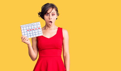 Beautiful young woman with short hair holding heart calendar scared and amazed with open mouth for surprise, disbelief face