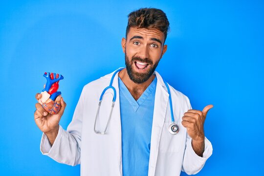 Young hispanic man wearing doctor uniform holding heart pointing thumb up to the side smiling happy with open mouth