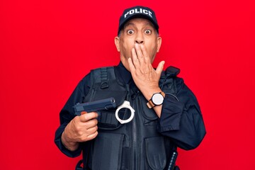 Senior man with grey hair wearing police uniform holding gun covering mouth with hand, shocked and afraid for mistake. surprised expression