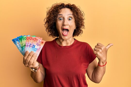 Beautiful middle age mature woman holding swiss franc banknotes pointing thumb up to the side smiling happy with open mouth
