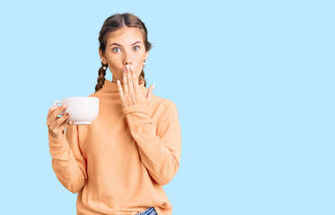 Beautiful caucasian woman with blonde hair drinking a coffee covering mouth with hand, shocked and afraid for mistake. surprised expression