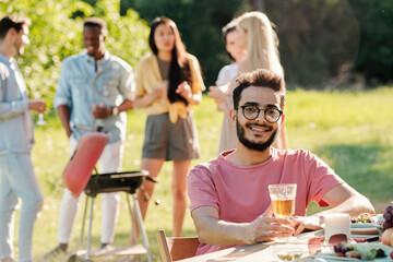 Young happy man with glass of wine looking at you while sitting by served table