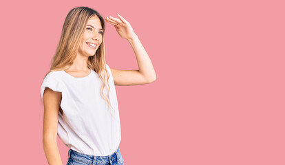 Beautiful caucasian woman with blonde hair wearing casual white tshirt very happy and smiling looking far away with hand over head. searching concept.