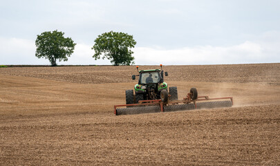 Obraz premium Farmer ploughing a field with his tractor equipped with a roll and press to prepare the soil before planting. Two trees isolated in the background. 