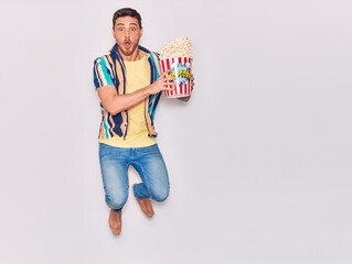 Young handsome hispanic man surprised with open mouth. Holding bucket of popcorn jumping over isolated white background.