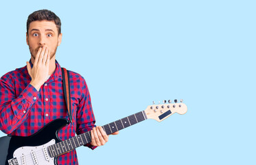 Handsome young man with bear playing electric guitar covering mouth with hand, shocked and afraid for mistake. surprised expression