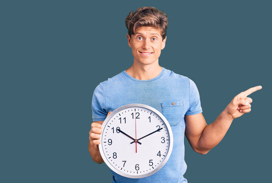 Young handsome man holding big clock smiling happy pointing with hand and finger to the side