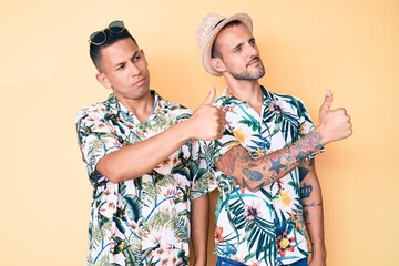 Young gay couple of two men wearing summer hat and hawaiian shirt looking proud, smiling doing thumbs up gesture to the side