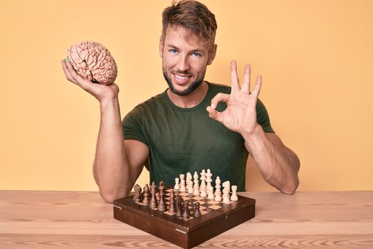 Young caucasian man holding brain while playing chess doing ok sign with fingers, smiling friendly gesturing excellent symbol