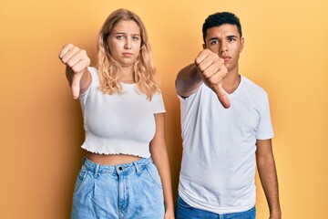 Young interracial couple wearing casual white tshirt looking unhappy and angry showing rejection and negative with thumbs down gesture. bad expression.