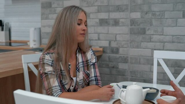 This video shows a girl sitting at the kitchen table. She listens to her interlocutor and drinks coffee.