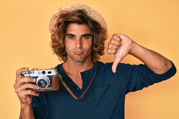 Young hispanic man wearing hat holding vintage camera with angry face, negative sign showing dislike with thumbs down, rejection concept