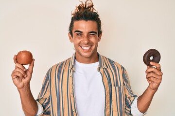 Young hispanic man holding red apple and donut winking looking at the camera with sexy expression,...