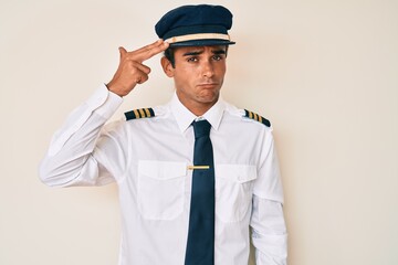 Young hispanic man wearing airplane pilot uniform shooting and killing oneself pointing hand and fingers to head like gun, suicide gesture.