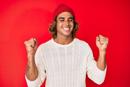 Young hispanic man wearing wool sweater and winter hat very happy and excited doing winner gesture with arms raised, smiling and screaming for success. celebration concept.
