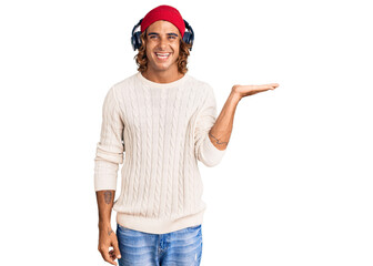 Young hispanic man listening to music using headphones smiling cheerful presenting and pointing with palm of hand looking at the camera.