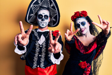 Young couple wearing mexican day of the dead costume over yellow smiling with tongue out showing fingers of both hands doing victory sign. number two.