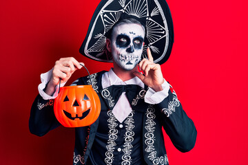 Young man wearing mexican day of the dead costume holding pumpkin serious face thinking about question with hand on chin, thoughtful about confusing idea