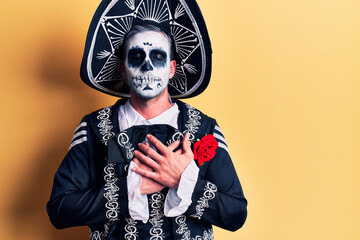 Young man wearing mexican day of the dead costume over yellow smiling with hands on chest with closed eyes and grateful gesture on face. health concept.